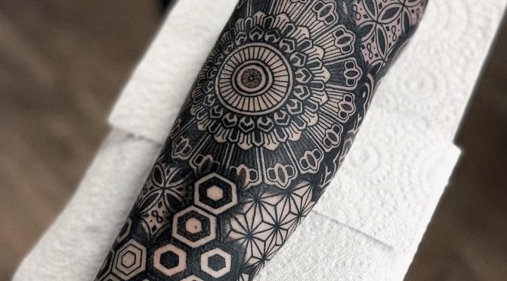 Black geometric forearm tattoo combining a circular mandala and repeating patterns
